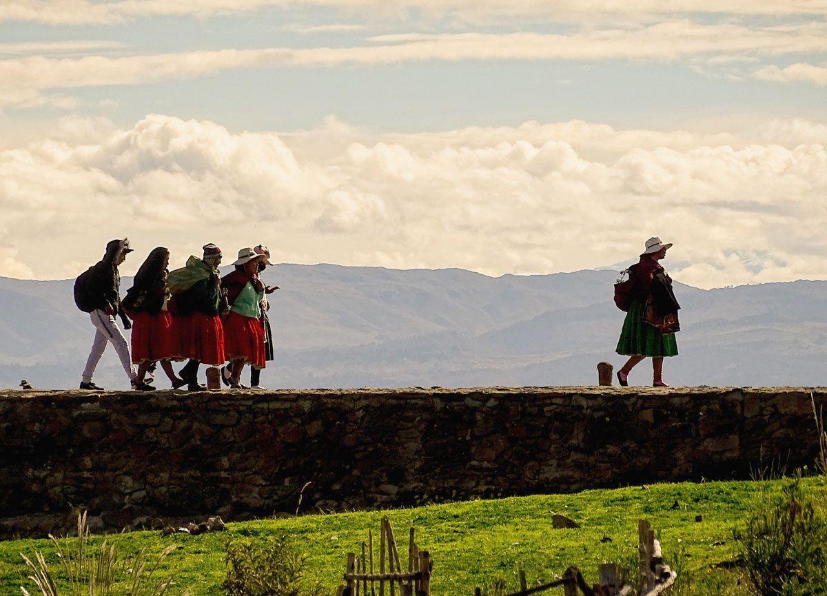 Six people walking on top of a stone wall with one more  person walking in front. Behind them is the Andes mountain range