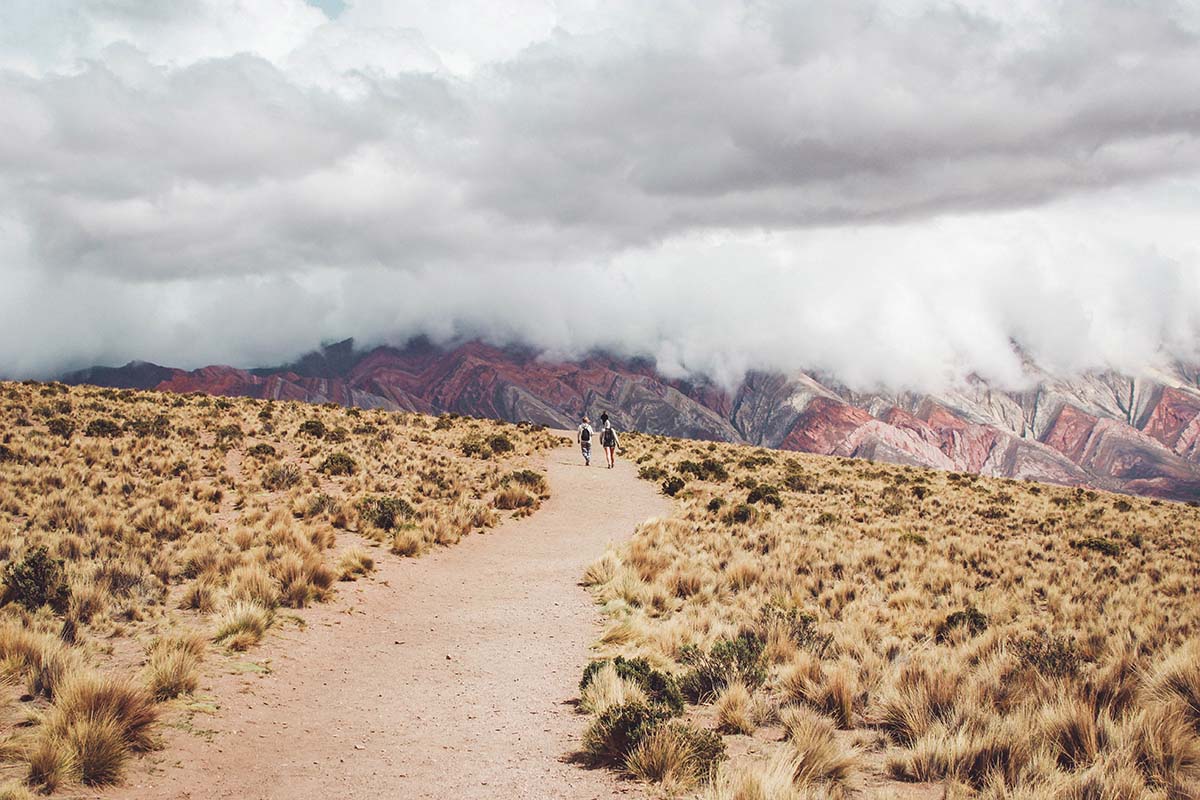Two hikers traverse a barren landscape and trail with colorful mountains in Humahuaca, Argentina.