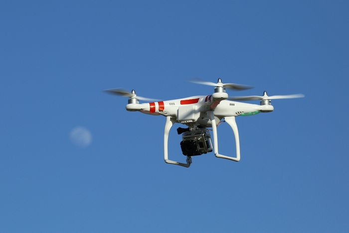 A white drone with red accents and a black camera flying in blue skies. A faint moon shape behind.