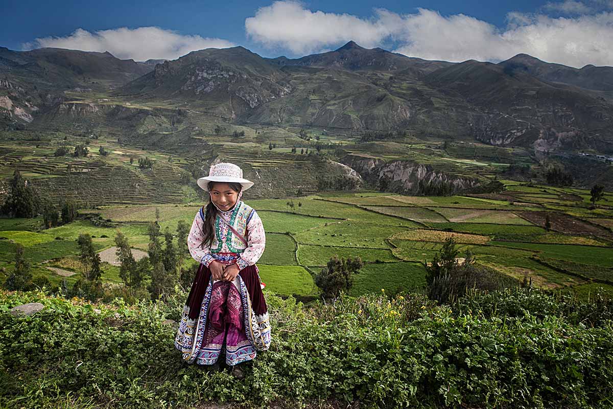 Young girl dressed in colorful traditional clothing with green terraces in Colca Canyon behind her.