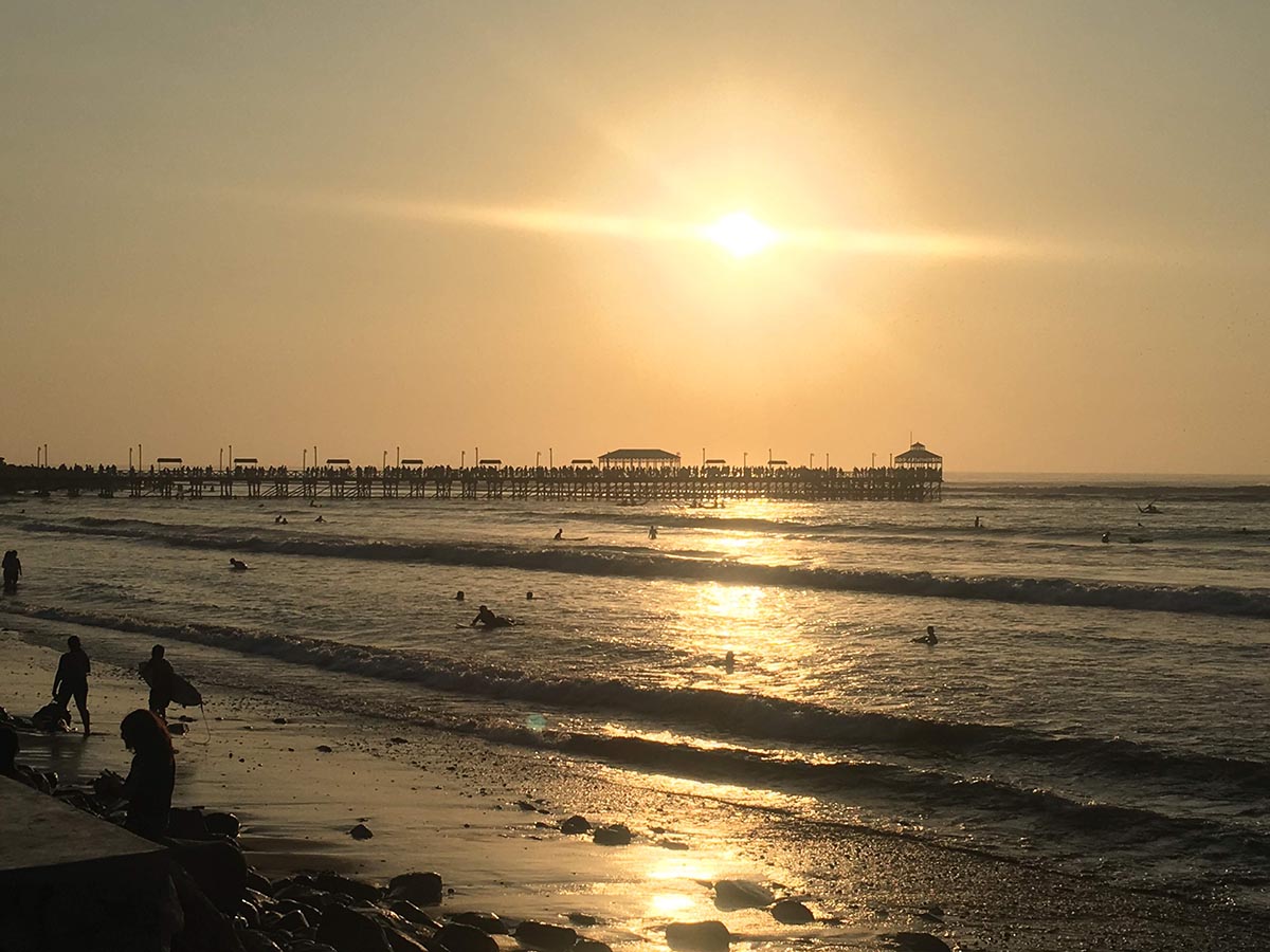 Sunset over a century-old pier in Huanchaco, Peru with people swimming in the low surf.