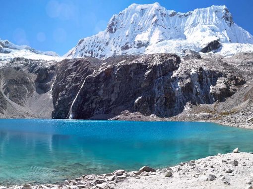 View of Laguna 69, a clear blue-green lake surrounded by snow-capped mountains and clear blue skies