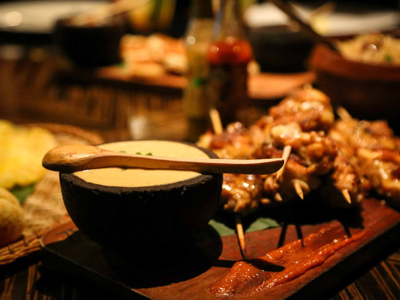 A black bowl with soup and a wooden spoon on top. Skewered meat blurred in the background.
