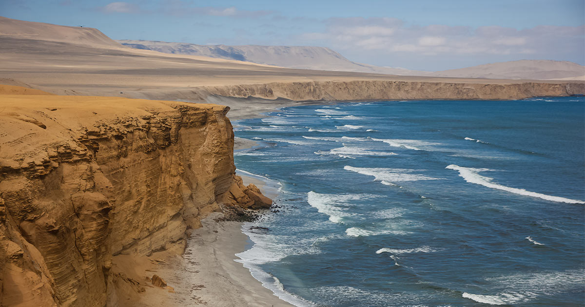 Waves crashing against the expansive desert cliff coast of the Paracas National Reserve in Peru.