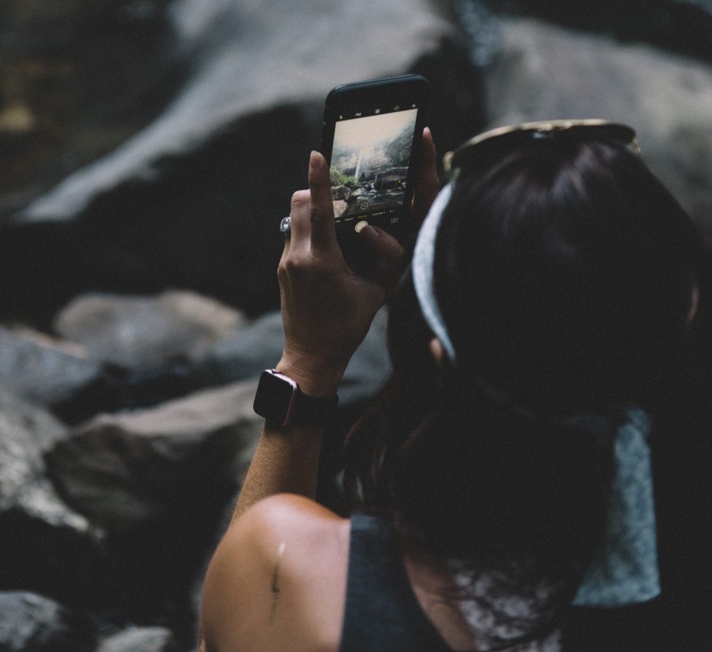 A woman holding a smartphone and taking a picture of a waterfall.