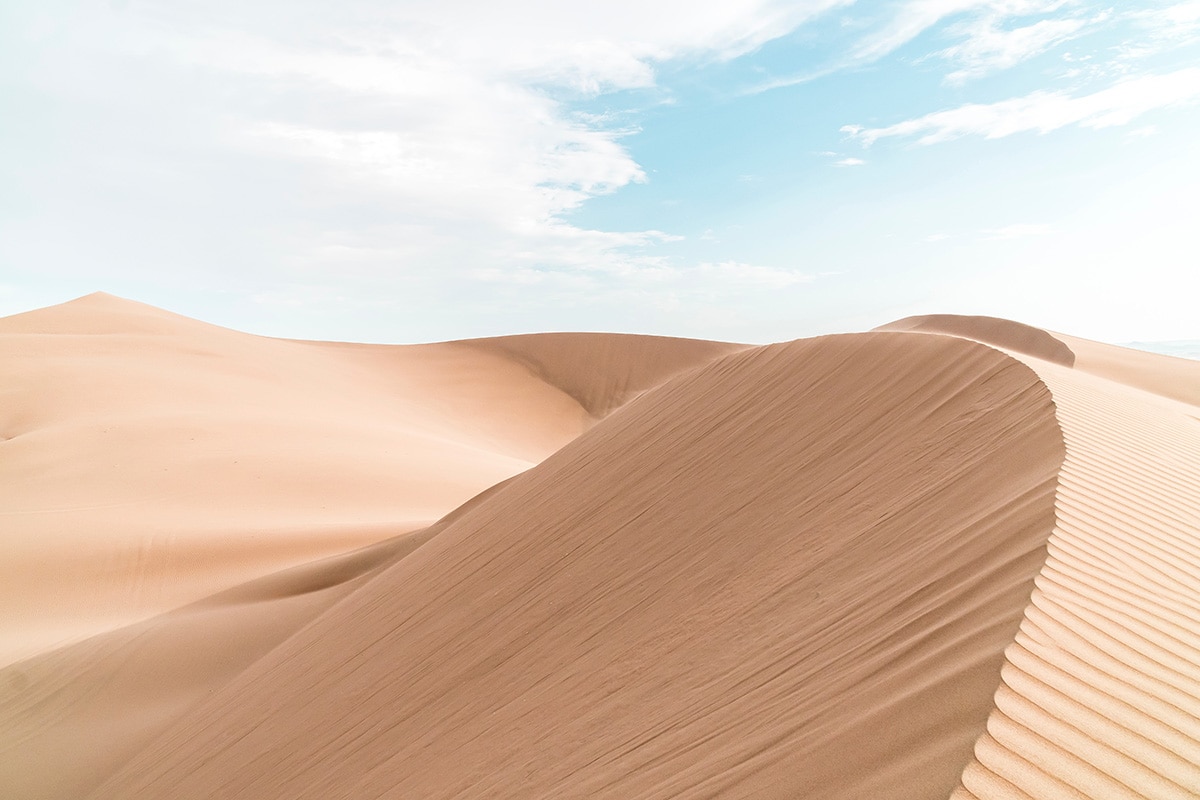 Sand dunes in the Peruvian desert with a cloudy blue sky backdrop.