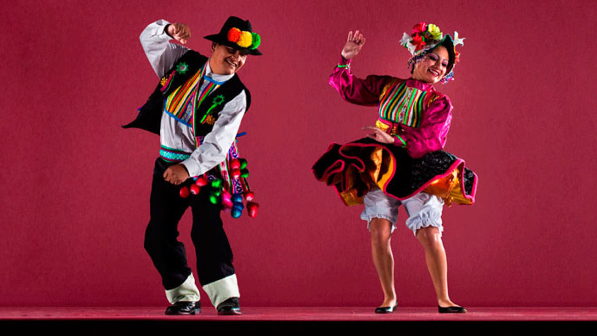 Two people dancing huayno in colorful attire with pink background.