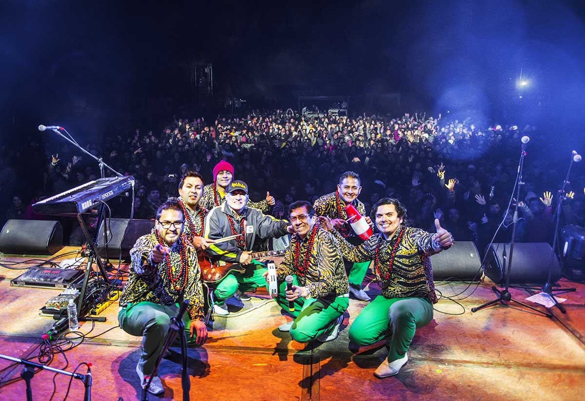 Seven performers of Los Mirlos smiling on stage with a live audience behind.