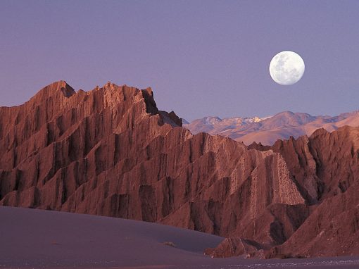 A full moon rises in a purple sky above mountainous rock formations in the Atacama Desert.