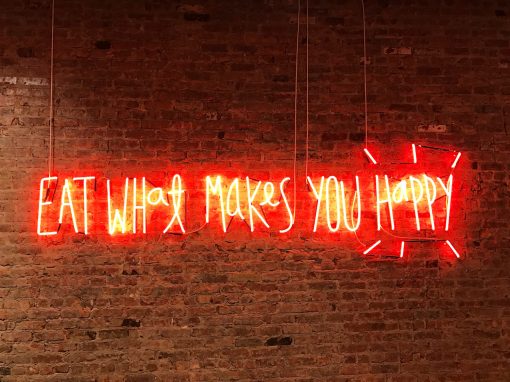 neon sign that says eat what makes you happy against a red brick wall