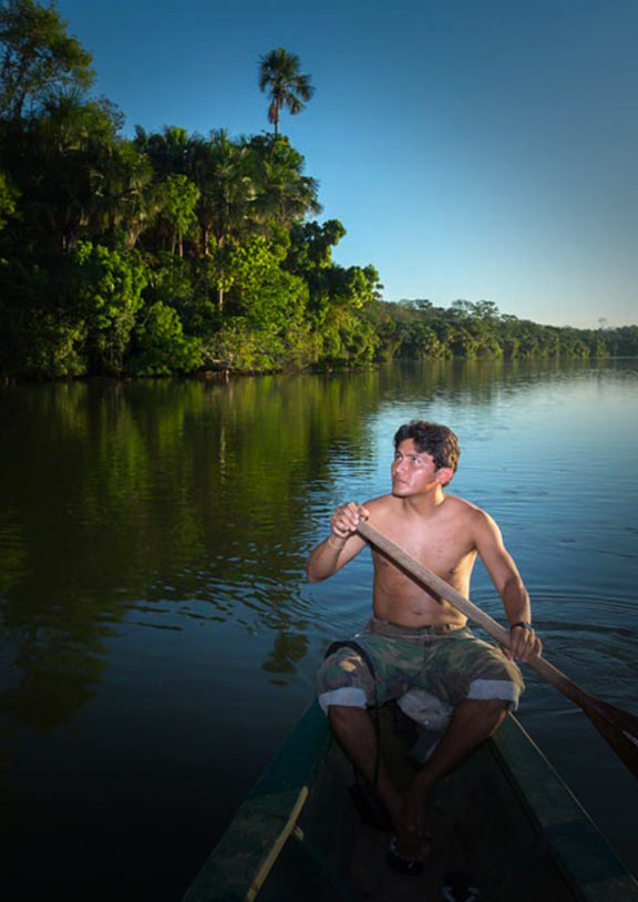 A man rows a boat on a river in the Amazon Rainforest with lush green trees growing on the shore