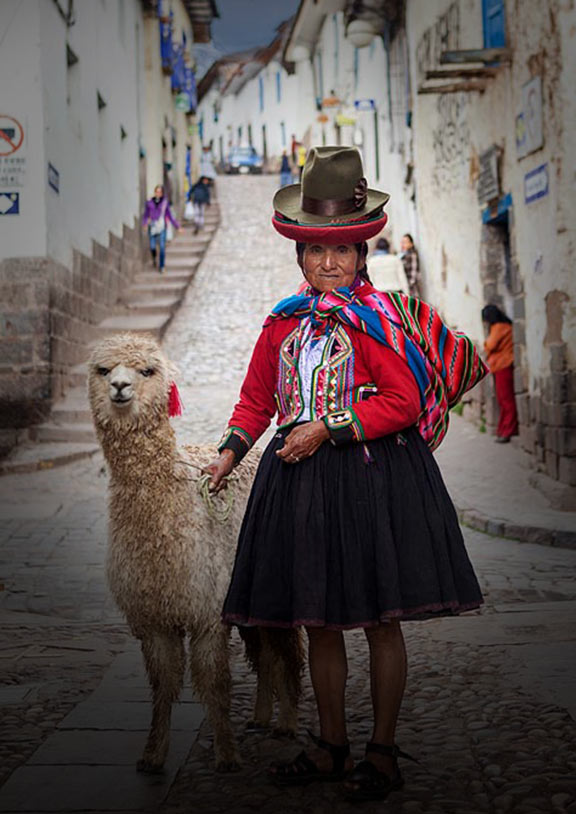 A woman wearing traditional Quechua clothing stands next to an alpaca with a narrow street behind