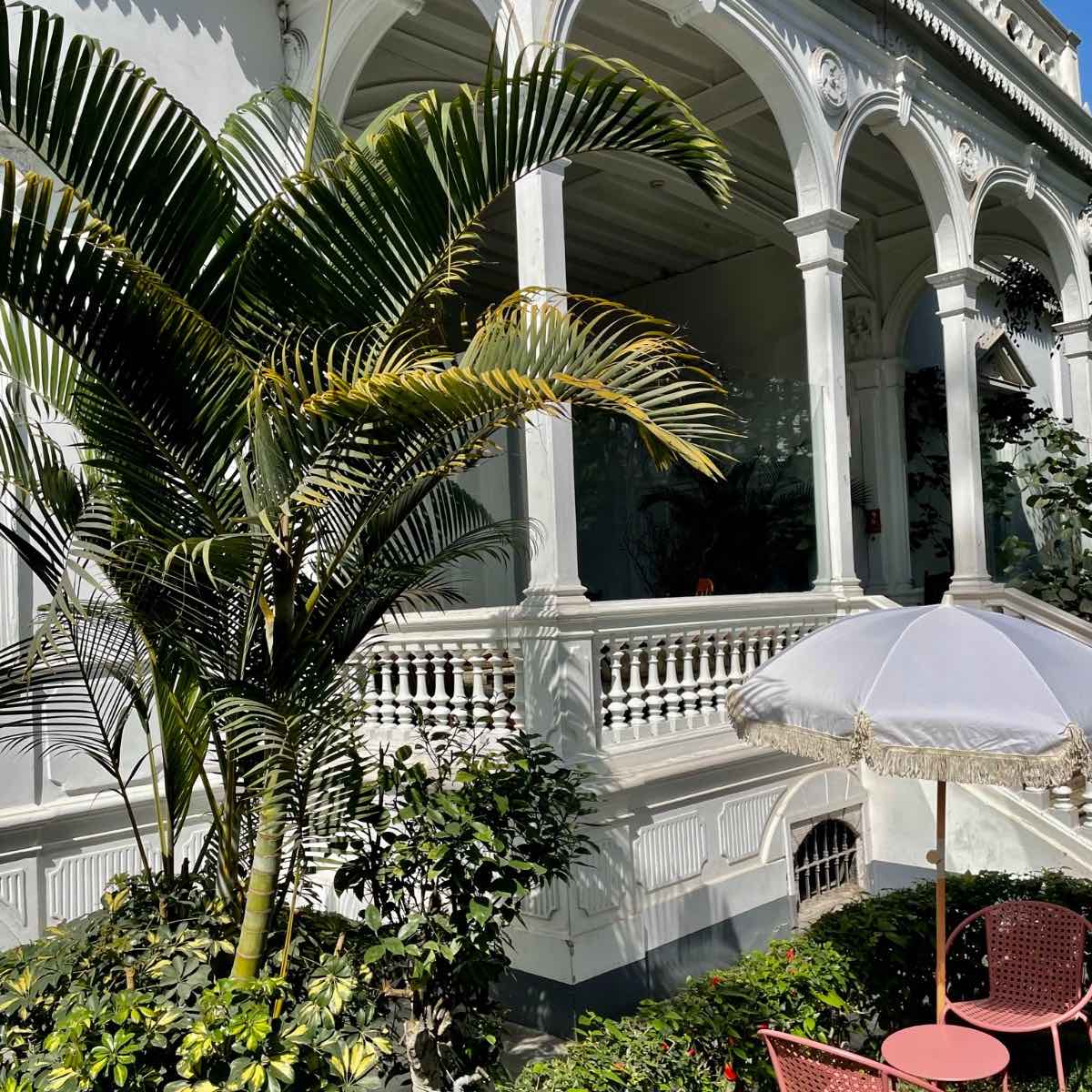 A white balcony with steps leads to a verdant garder where two pink chairs are laid out for visitors. 