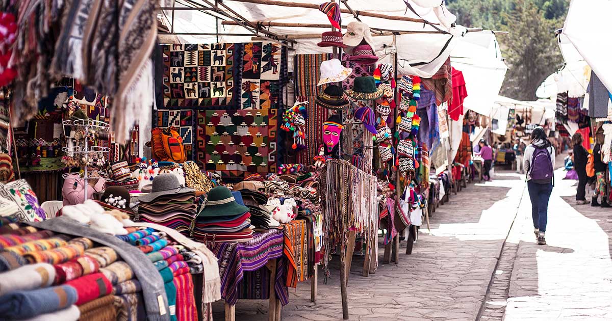 An outdoor Peruvian market. Stalls are selling alpaca scarves, hats, pompoms, and wall hangings.
