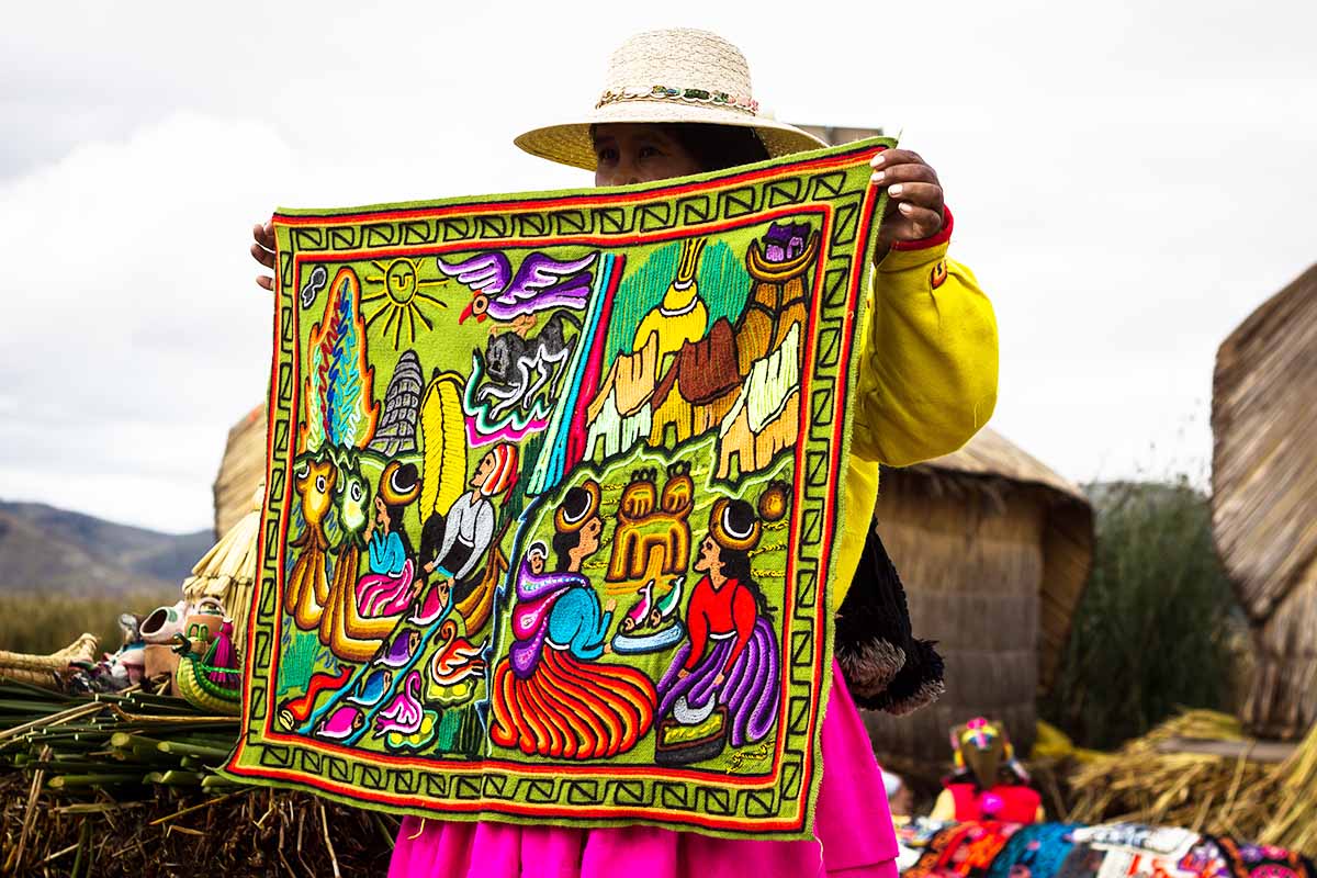 An Andean woman holds up a woven textile piece that shows scenes of Andean life.