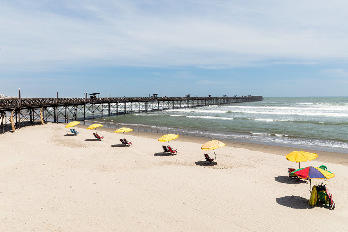 Light colored sand with a row of yellow beach umbrellas next to a pier leading into the ocean.