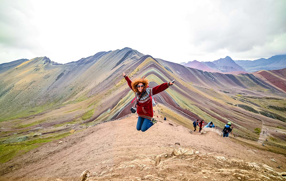A woman with bright red hair jumps for a photo in front of the Rainbow Mountain in Peru.