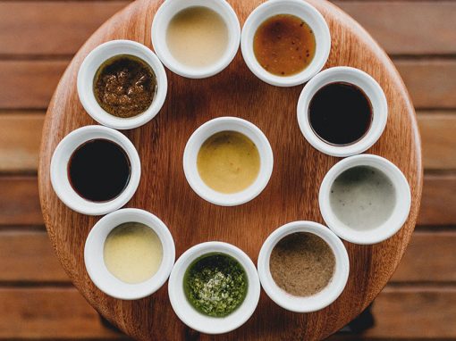 Nine white cups holding a variety of sauces arranged around a wood plate with wood table underneath.