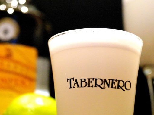 A cup of pisco sour labeled Tabernero with a bottle, some limes and another glass behind.